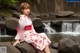 A woman in a red and white kimono sitting on a rock by a waterfall.